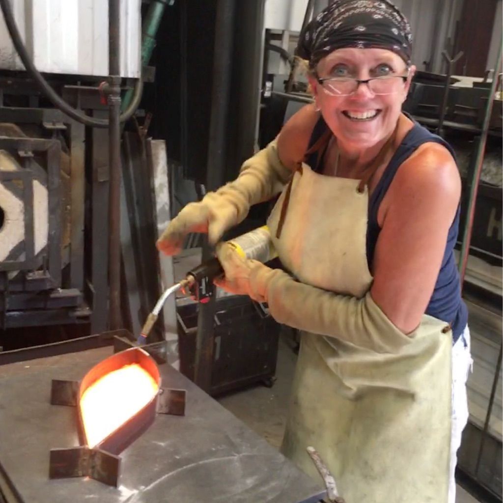 A woman wearing an apron and gloves is pouring molten metal into a ladle