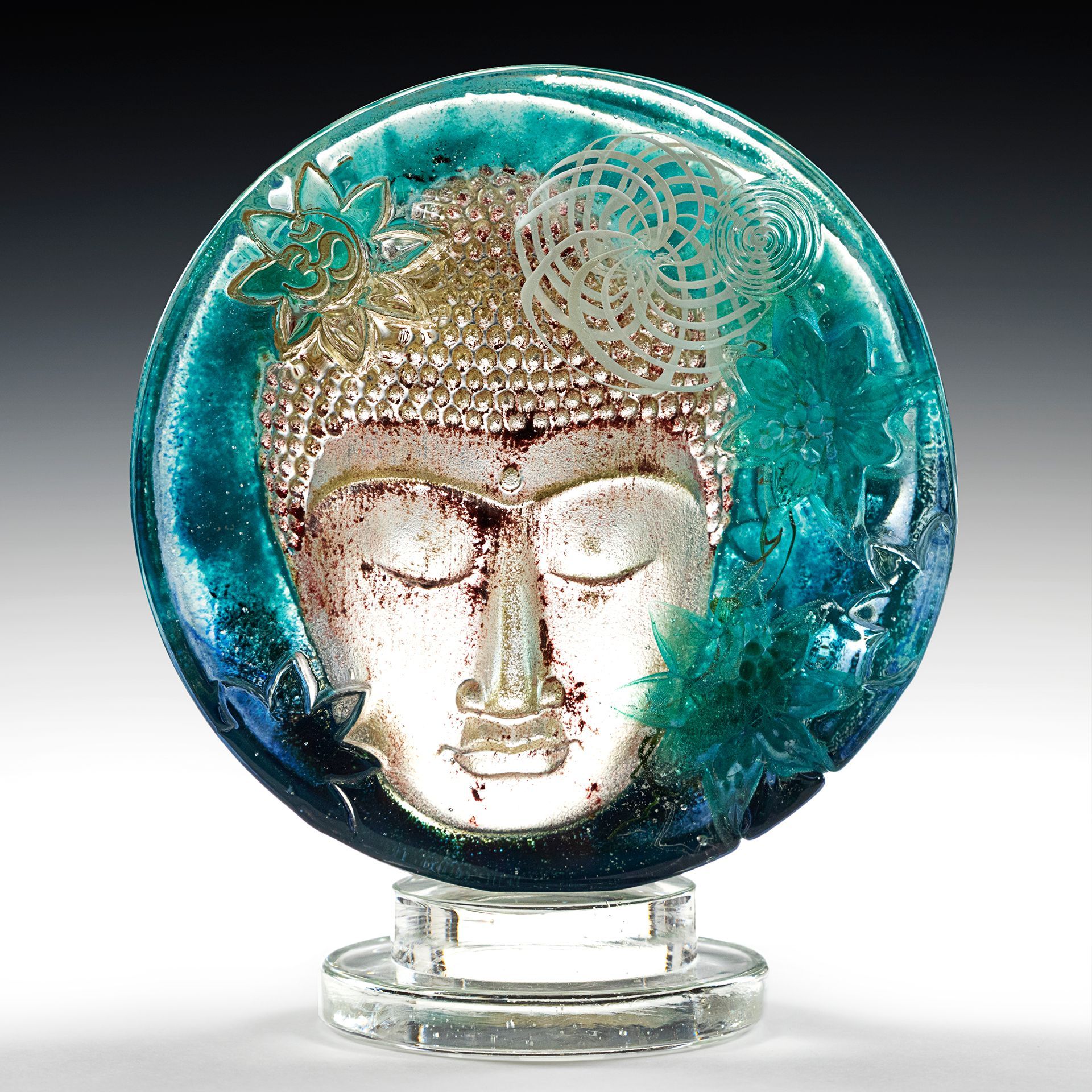 Glass sculpture of a serene Buddha face within a teal and gold circle, mounted on a clear stand.