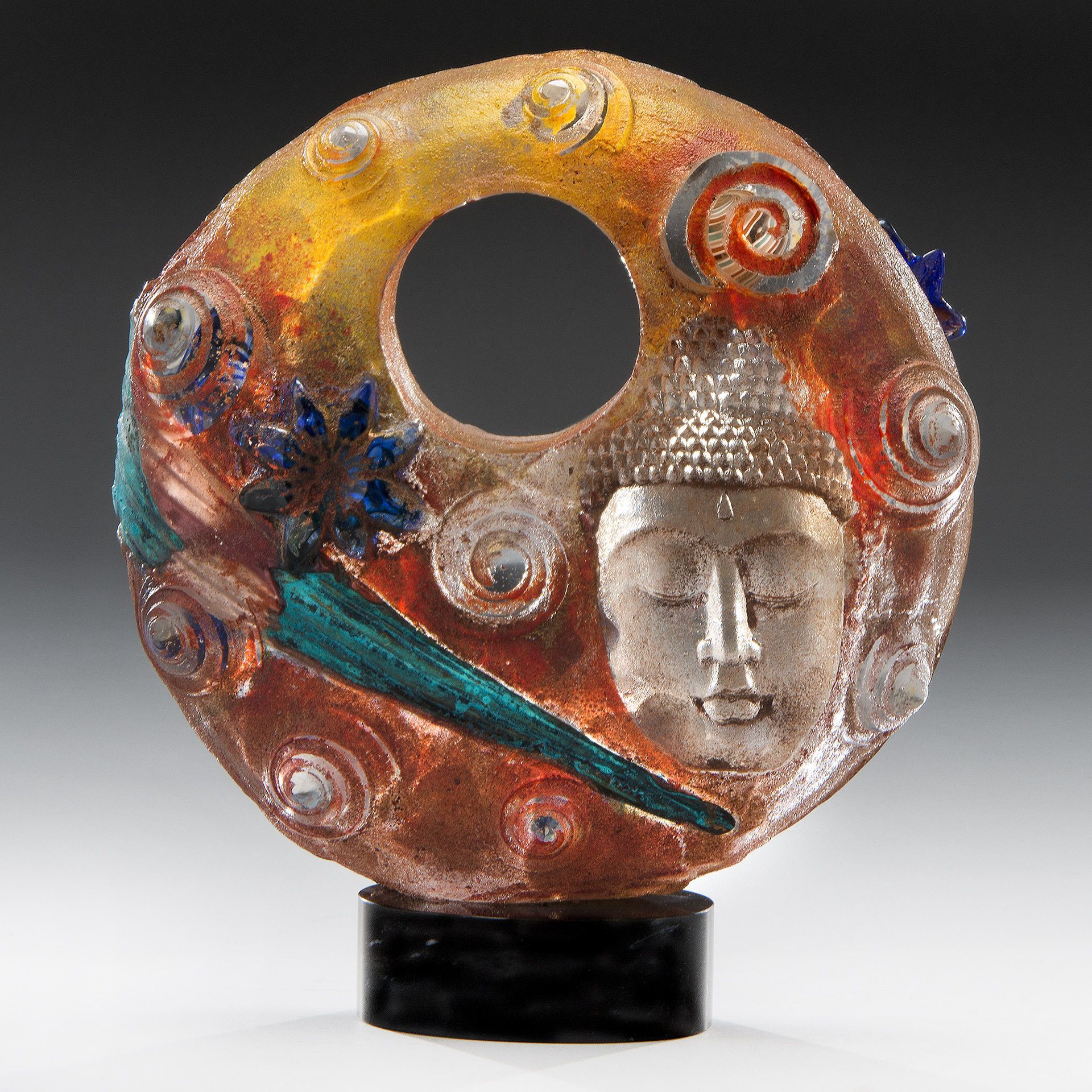 Glass sculpture of a Buddha face with a central circle and swirling colors; mounted on a black base.