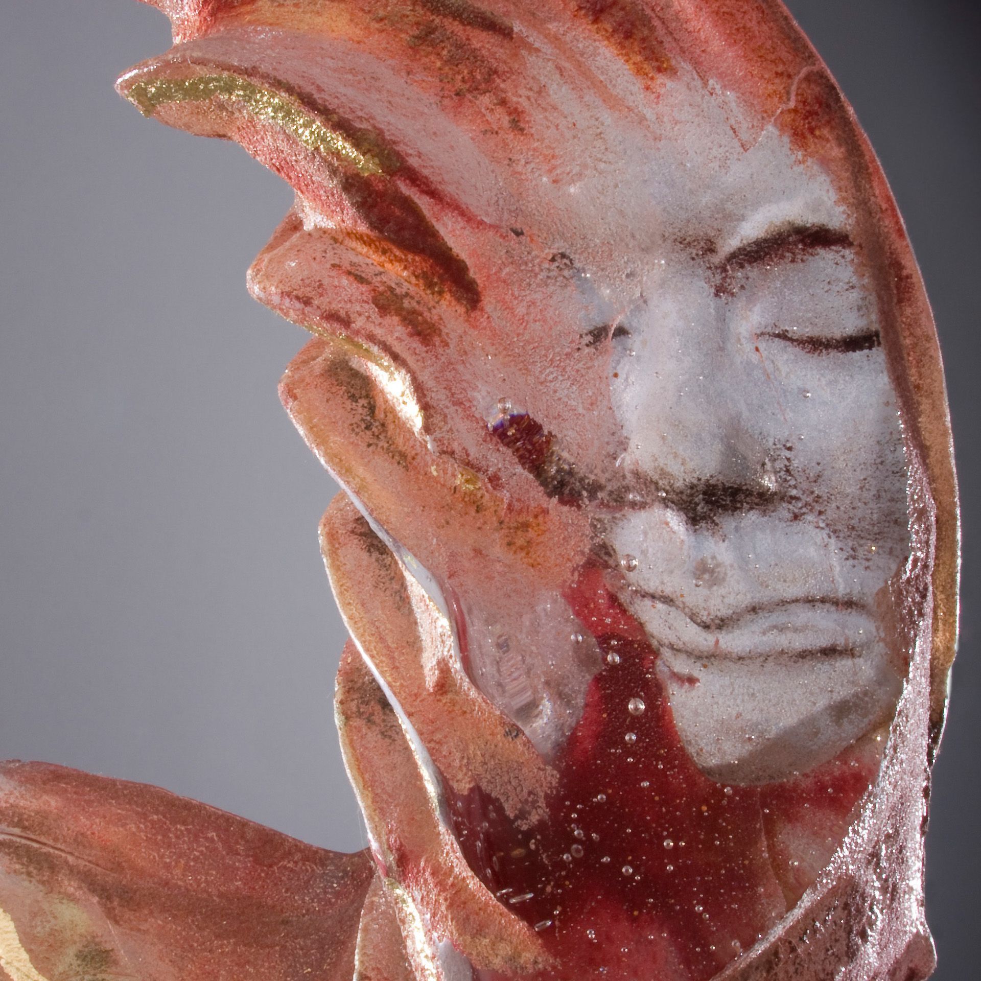 Glass sculpture of a serene face with closed eyes, hands, and flowing reddish, gold, and white colors.
