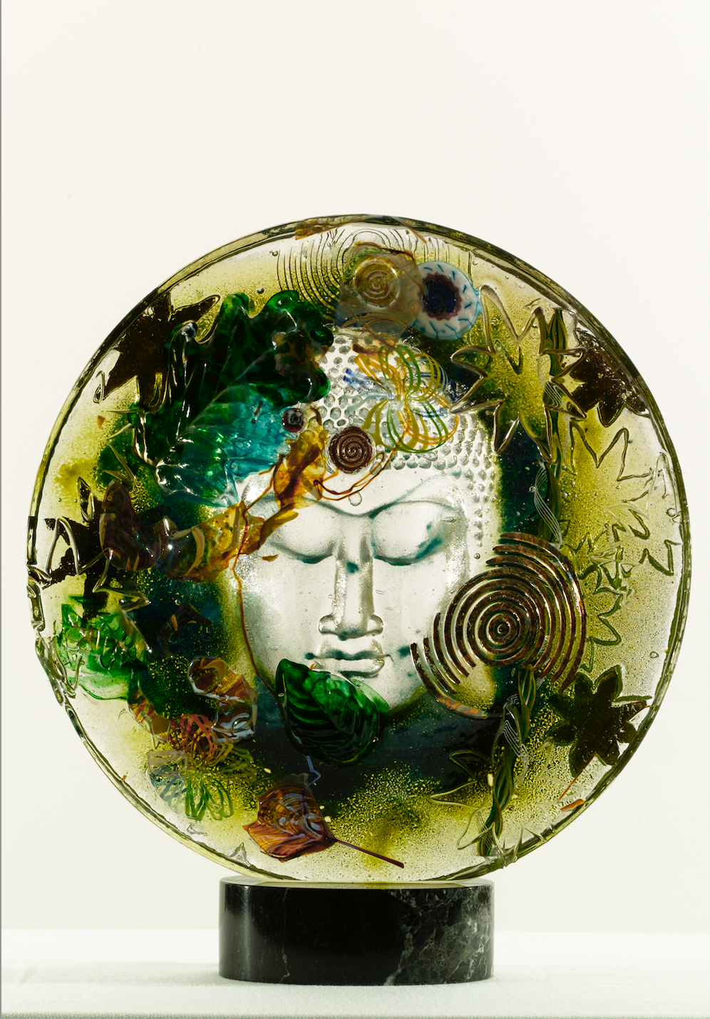Glass sculpture of Buddha's face, encircled by leaves and a spiral, mounted on a black base.
