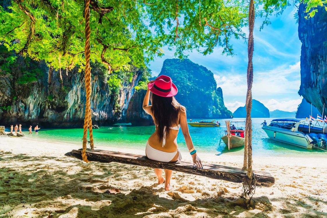 Mujer con sombrero rojo sentada en un columpio en una playa tropical con aguas turquesas y acantilados de piedra caliza.