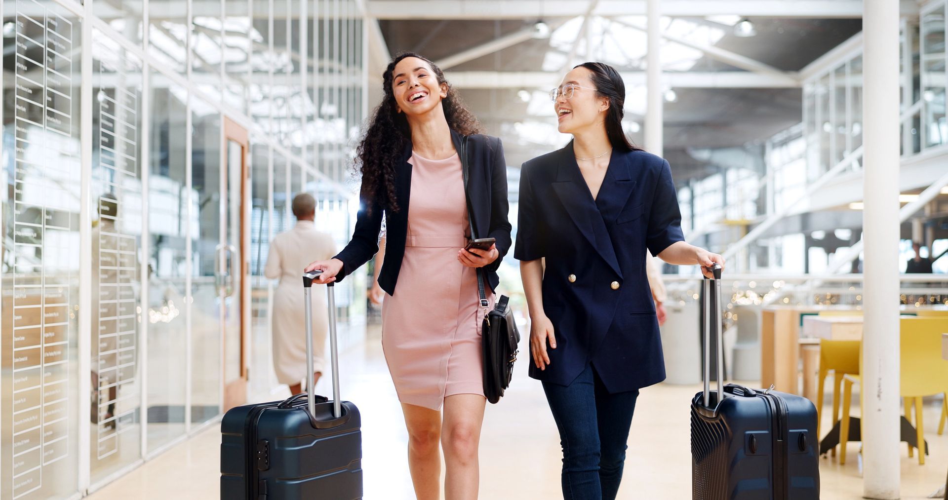 Dos viajeros caminan por una luminosa terminal de aeropuerto, cada uno arrastrando una maleta y sonriendo.