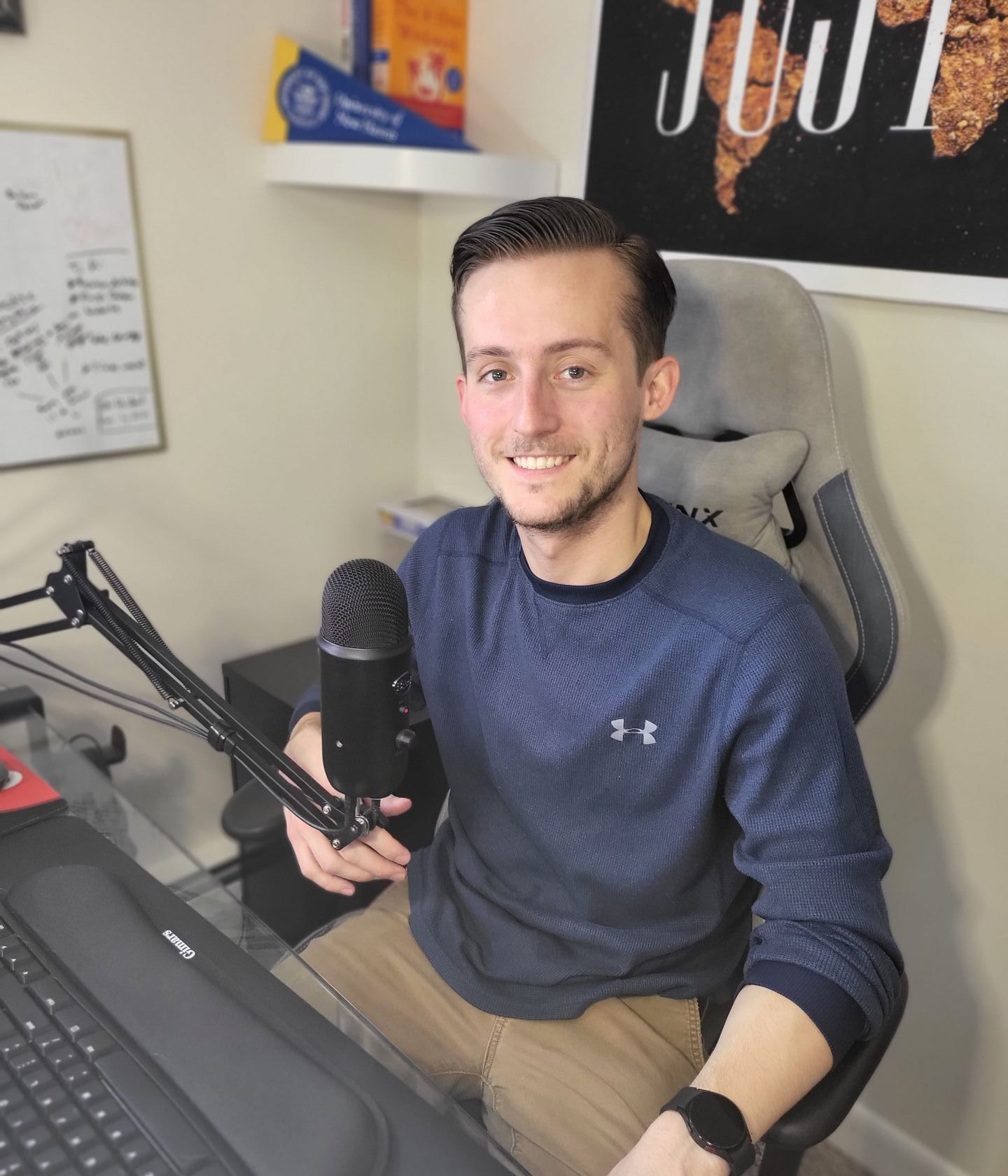 Person at a desk with a microphone and laptop in a home office, smiling at the camera.