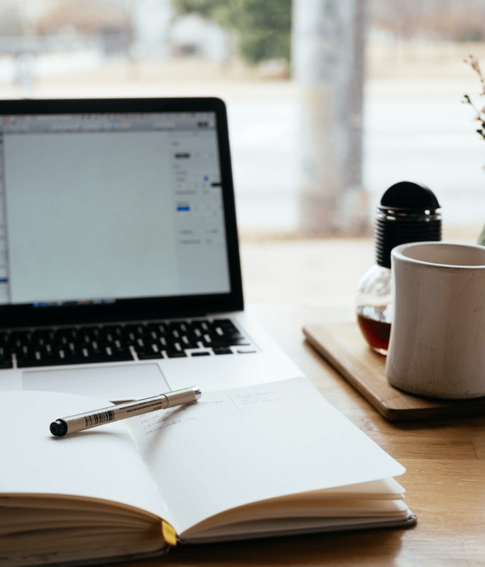 Notebook, pen, and laptop on a wooden desk beside a mug and tray in a bright workspace