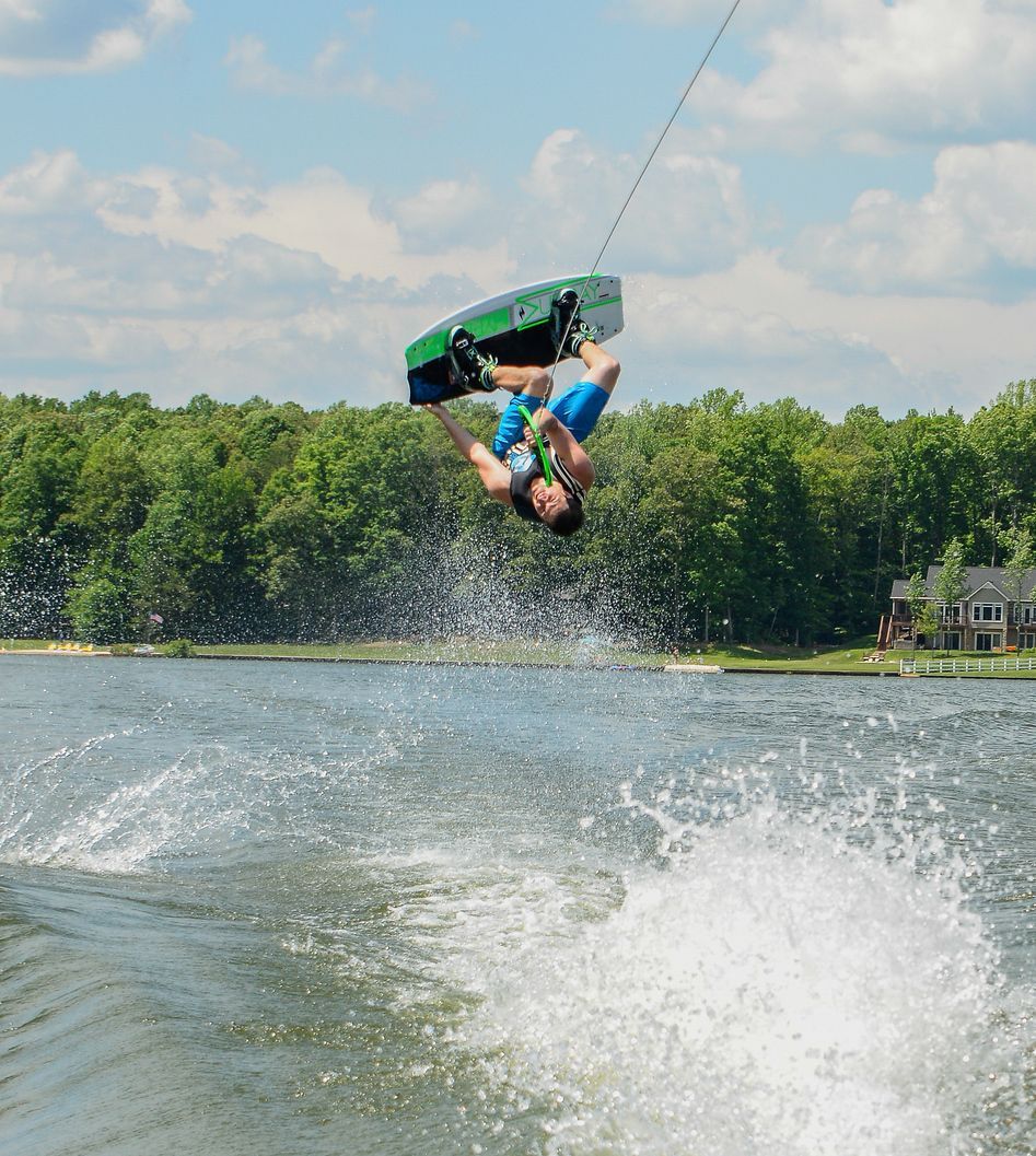 Wakeboarder flips midair above a lake, towing behind a boat with trees in the background.