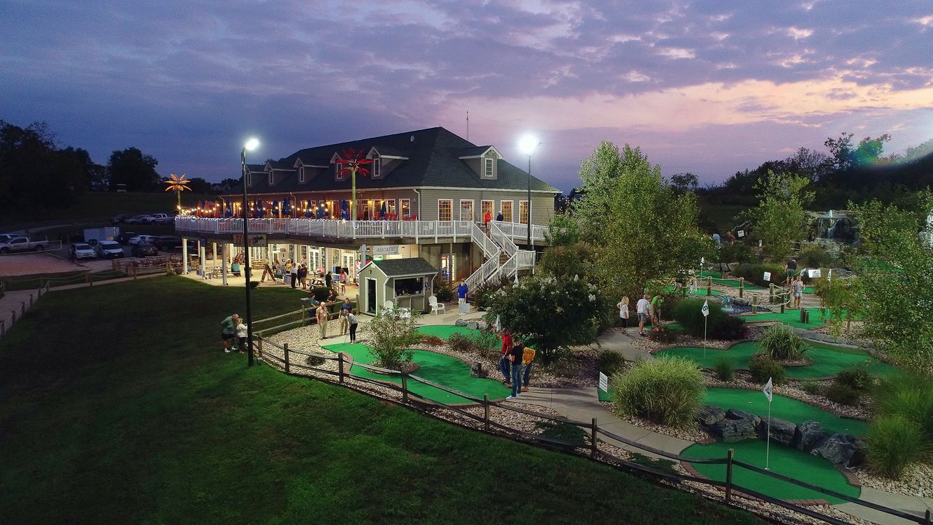 Lit outdoor restaurant and garden at dusk with green lawns and purple sunset sky