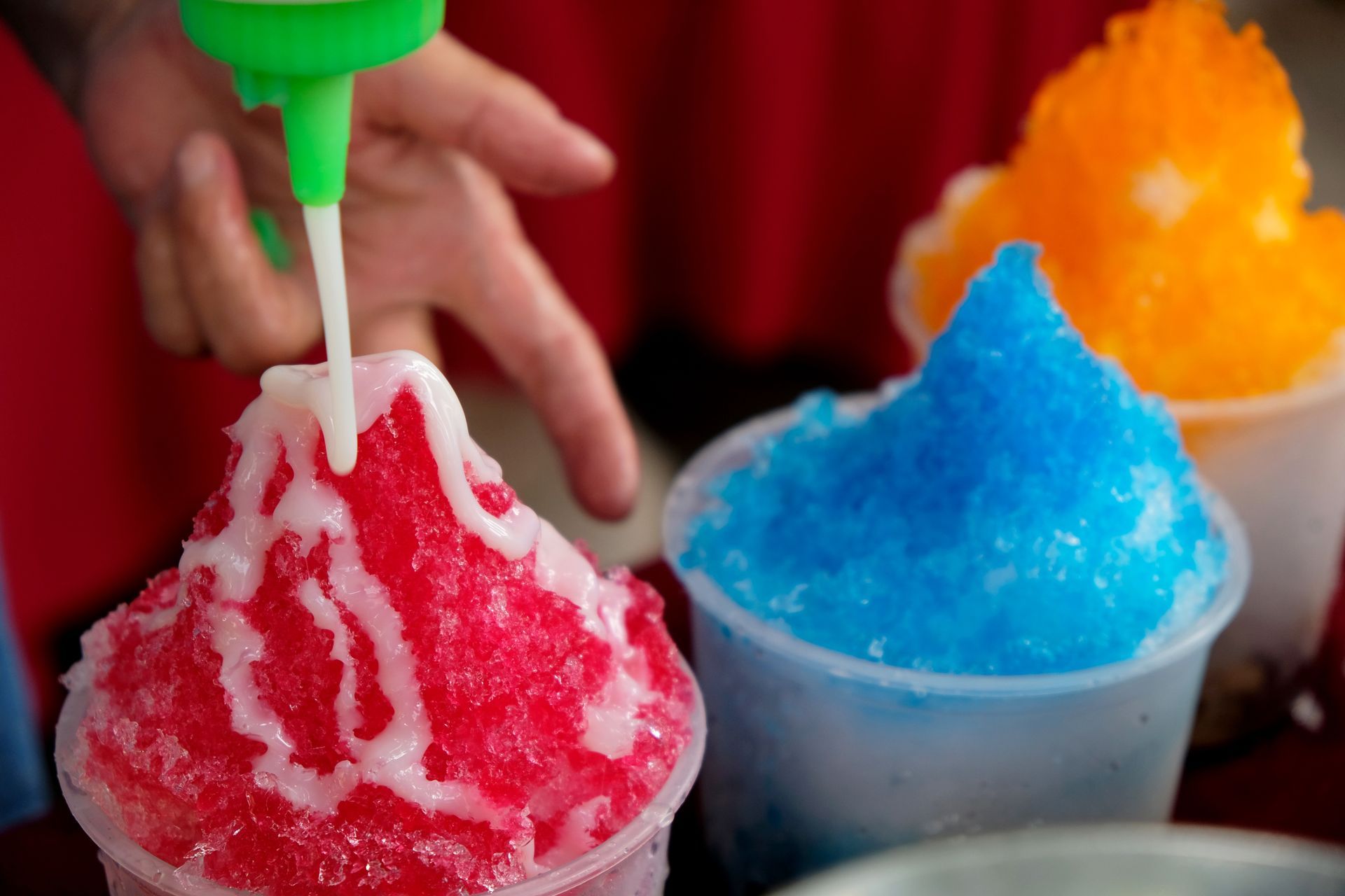 Shaved ice being topped with syrup: red in front, blue and orange behind.