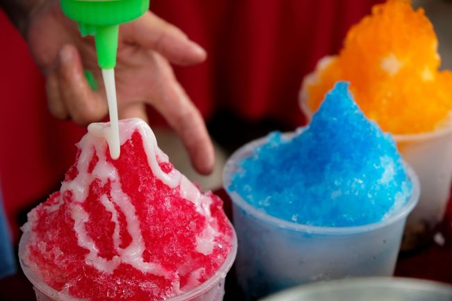 Shaved ice being topped with syrup: red in front, blue and orange behind.