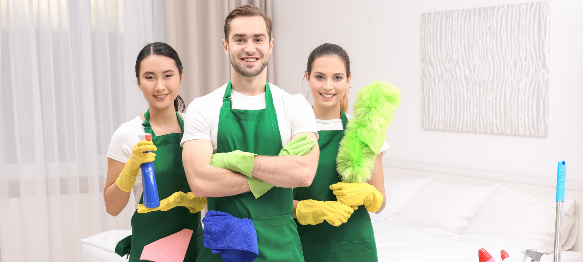 Three smiling professional cleaners wearing green aprons are holding the spray bottle and duster.