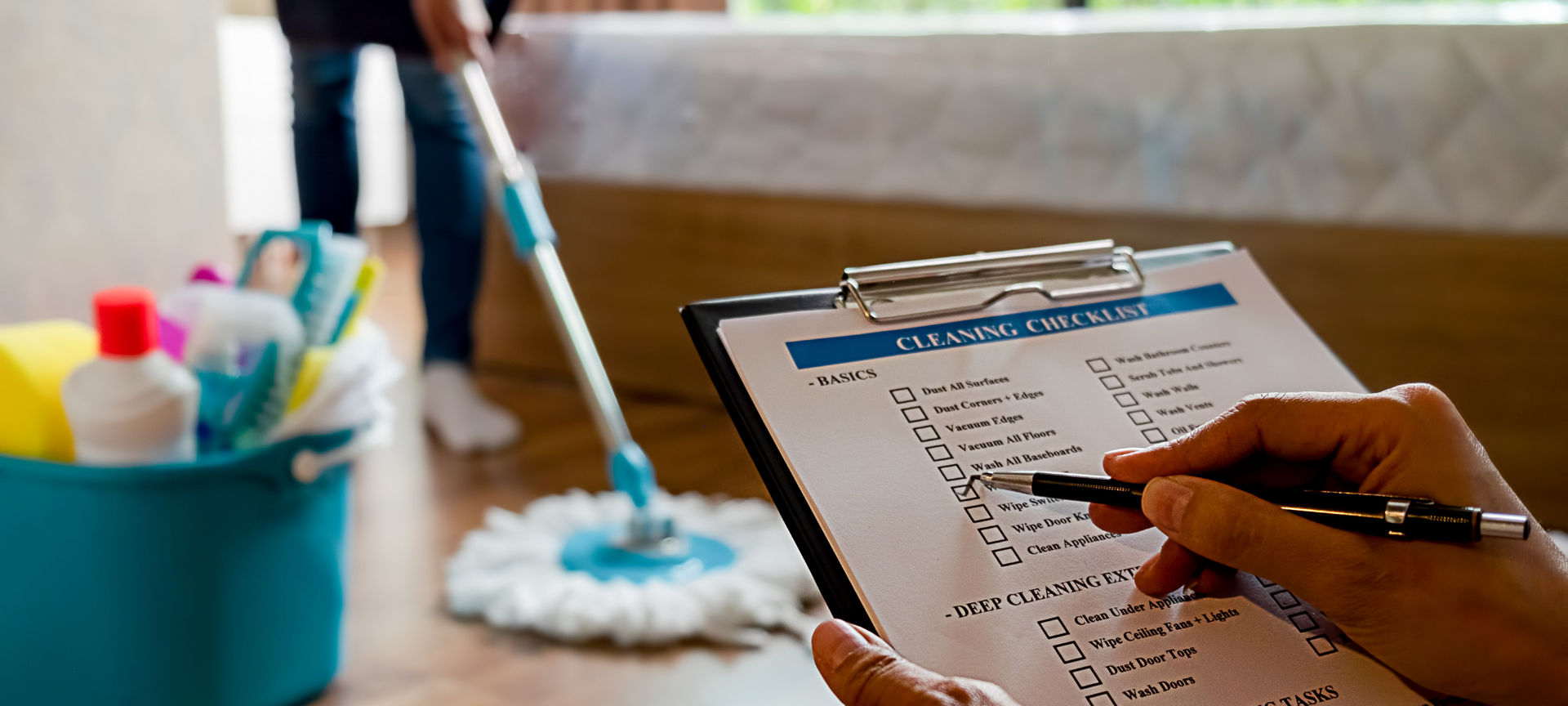 A close view of a hand confirming completion of items on a professional cleaning checklist while someone mops in the background.