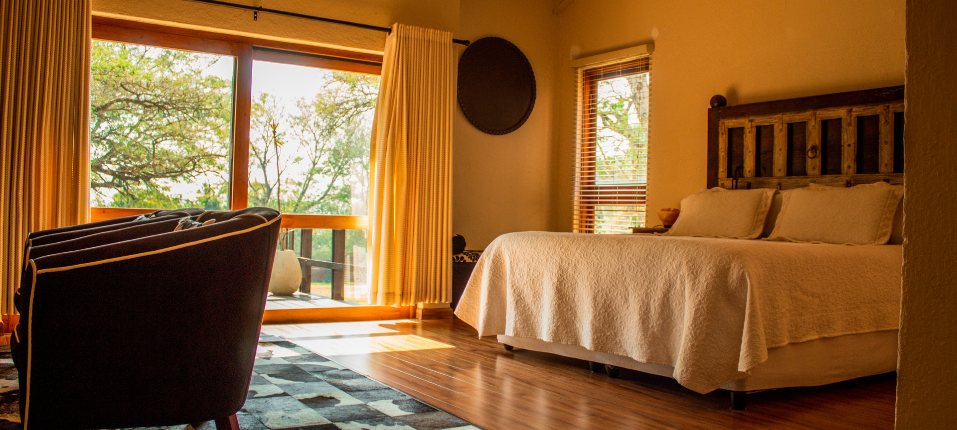 A rustic, warm bedroom has a big window with nature views and a bed topped with a white textured coverlet.