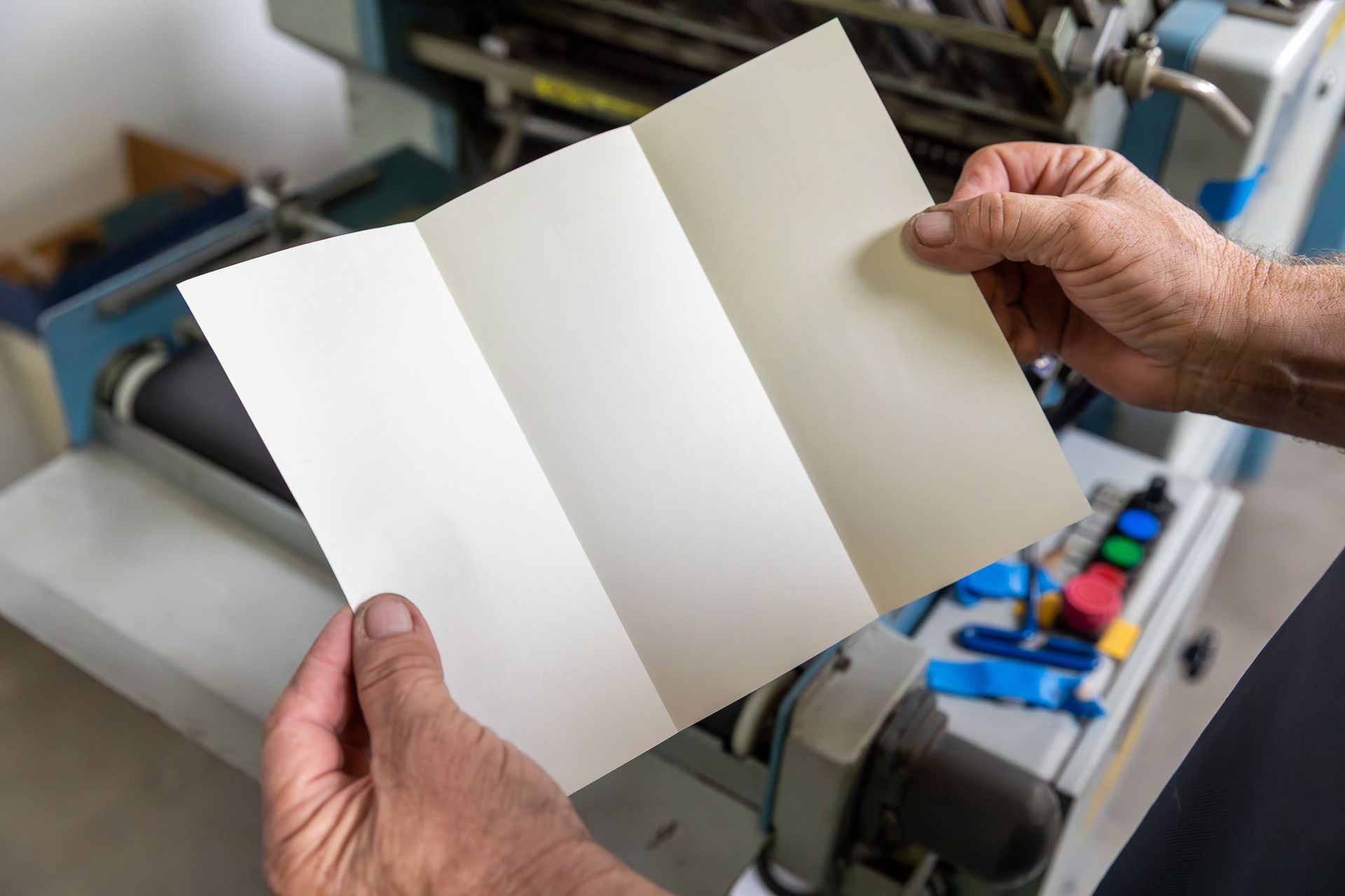 Person holding a tri-fold brochure, likely freshly printed, in front of a printing machine.