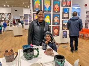 Woman and child at art exhibit, smiling. Pottery on display, artwork on walls.