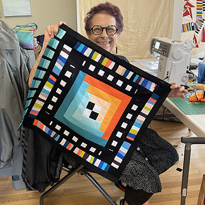 A person smiles while holding up a square quilt featuring a vibrant orange-to-teal gradient log cabin pattern with black.