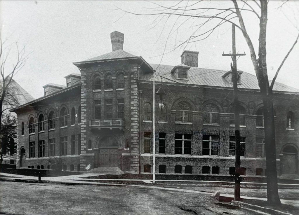 Historic brick building with arched windows and a central tower, on a street corner, with utility pole in front.