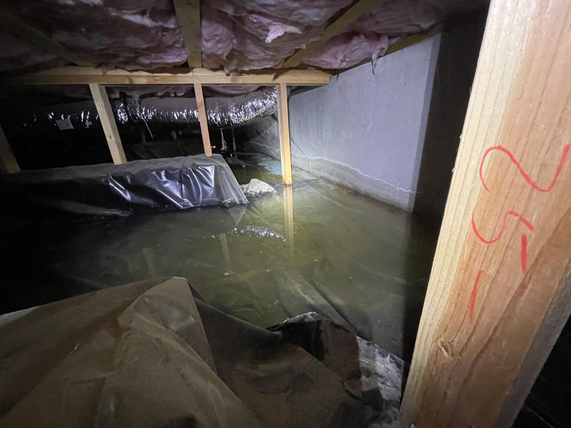 Flooded crawl space with standing water, exposed beams, and plastic-covered insulation under a house