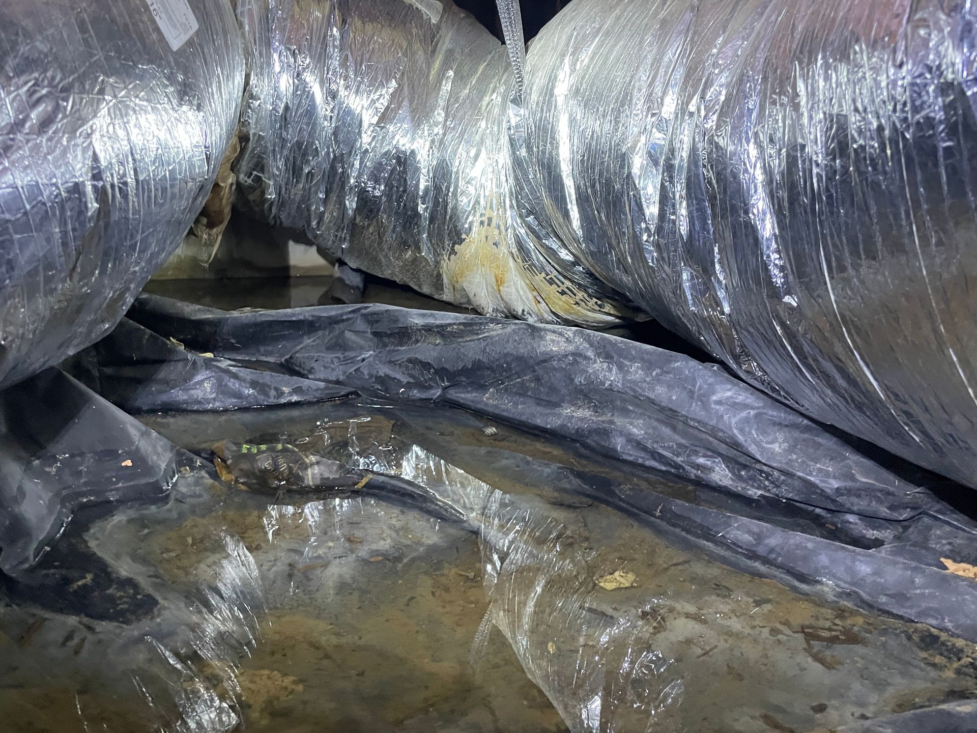 Black plastic ducting and foil-wrapped pipes above a wet, dirty floor in a cramped industrial crawlspace