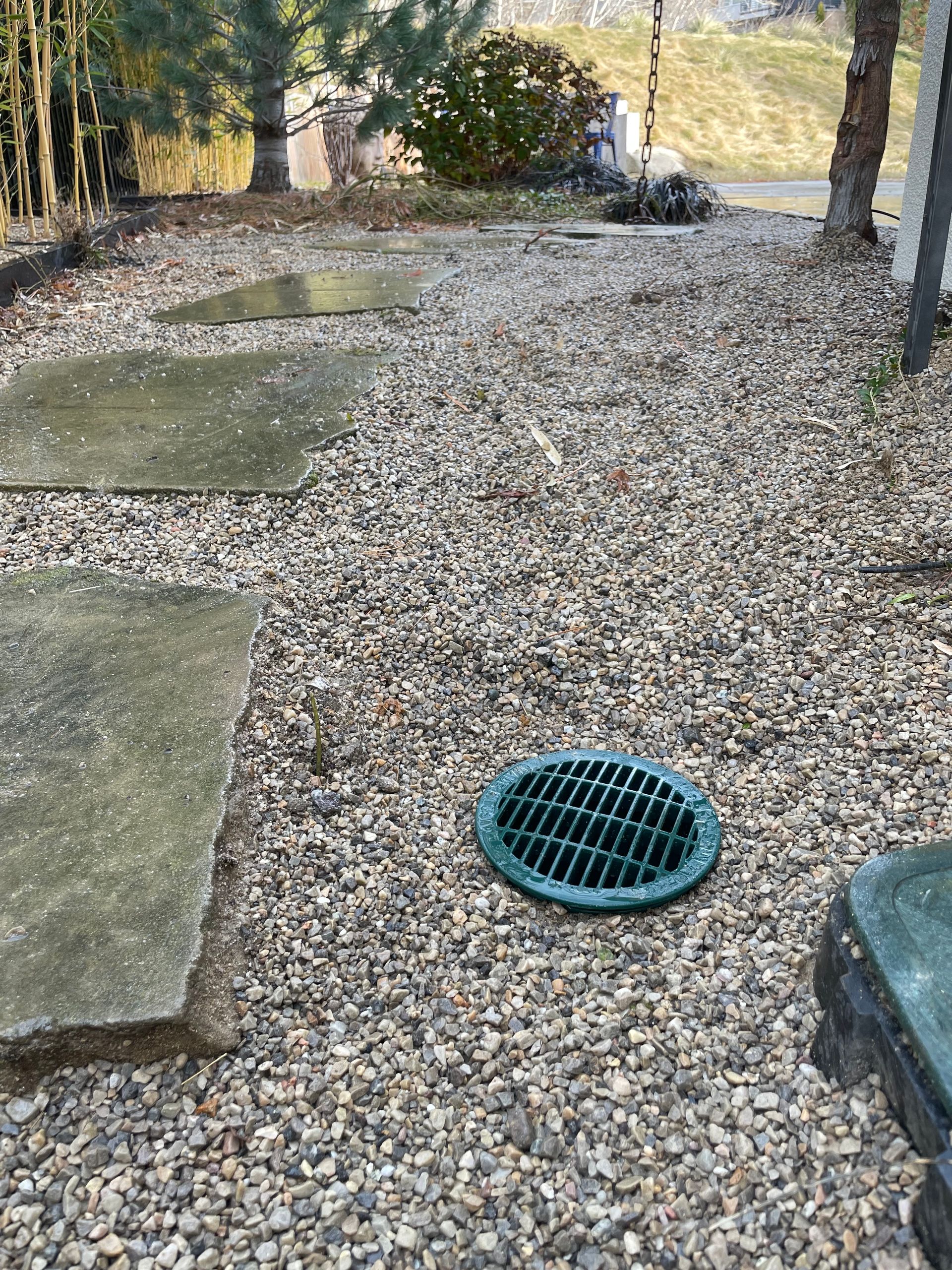 Gravel path with green drain cover beside a stone walkway, bordered by trees in a park-like setting