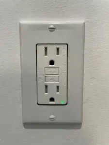 White GFCI electrical outlet on a light gray wall, with a green indicator light.