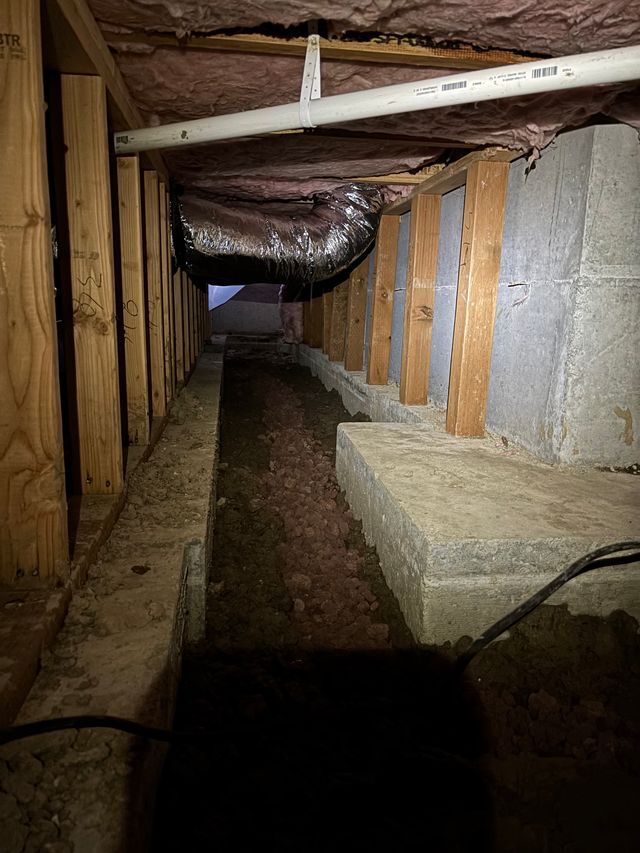 Crawlspace with wooden framing, concrete blocks, ducts, and dirt floor under a house.
