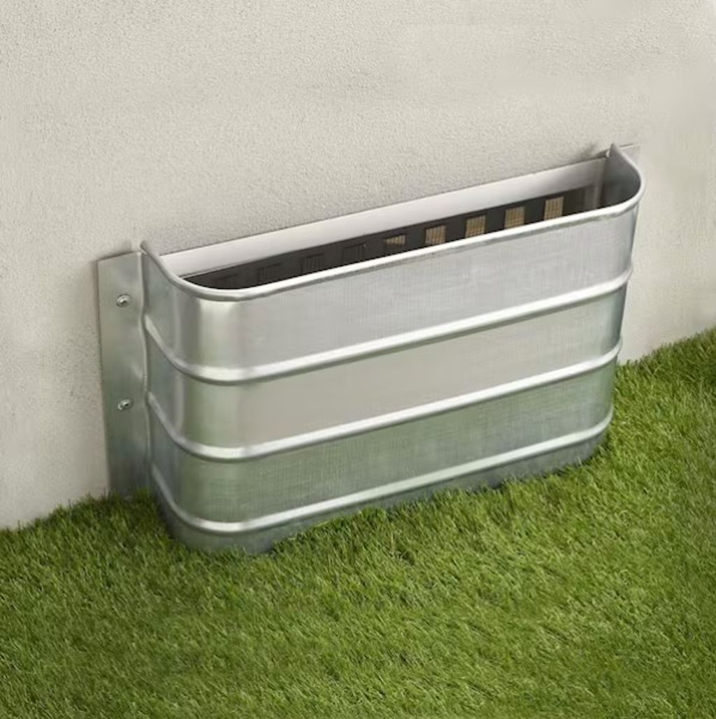 Metal well vent mounted against a beige wall above green grass