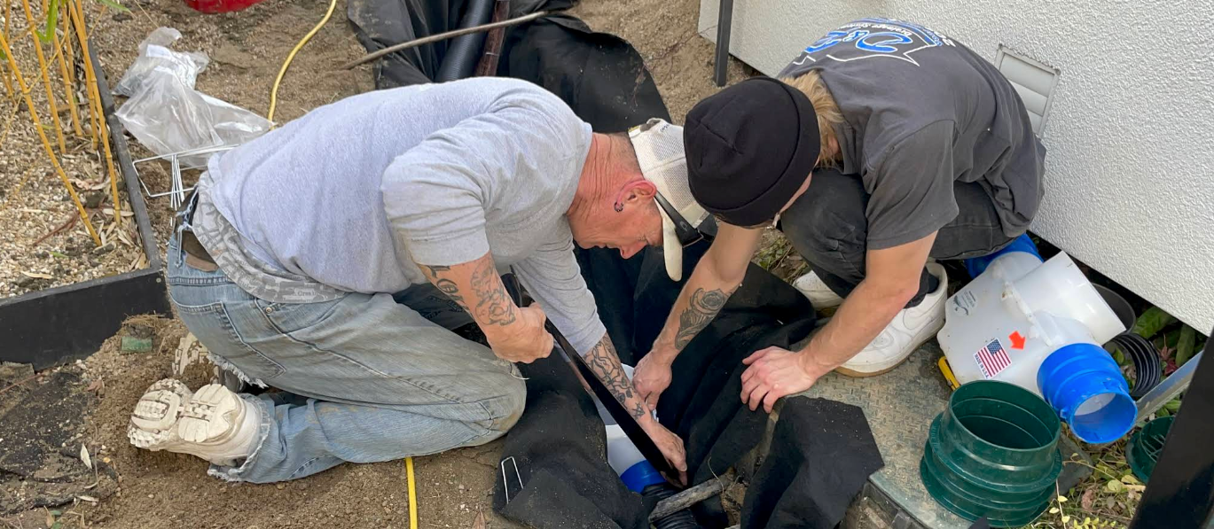 Two people kneel over a black dog on dirt beside a blue bucket and scattered supplies.