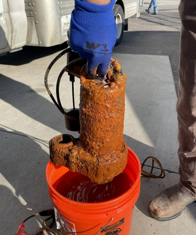 Rusty pipe segment being lifted from an orange bucket by a gloved worker outdoors