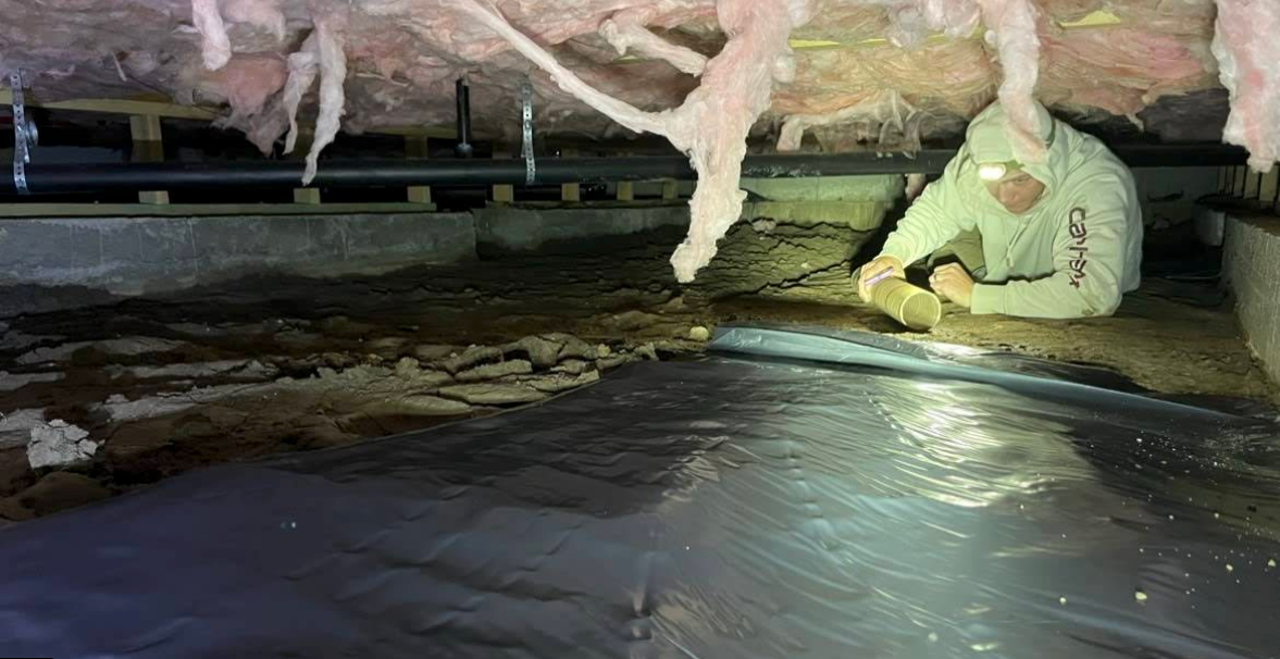 Person kneeling under a crawlspace, inspecting insulation and plastic sheeting with a flashlight.