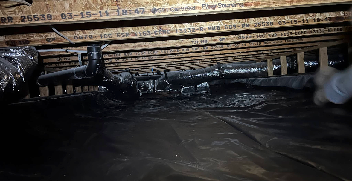 Inside a crawlspace, a dark plastic vapor barrier covers the floor beneath wooden beams and pipes.