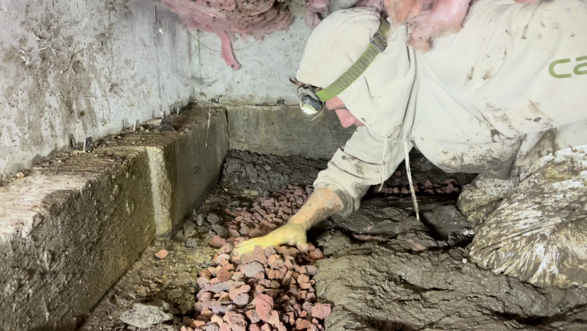 Worker in protective gear kneeling in a muddy trench, clearing debris beside a concrete wall.