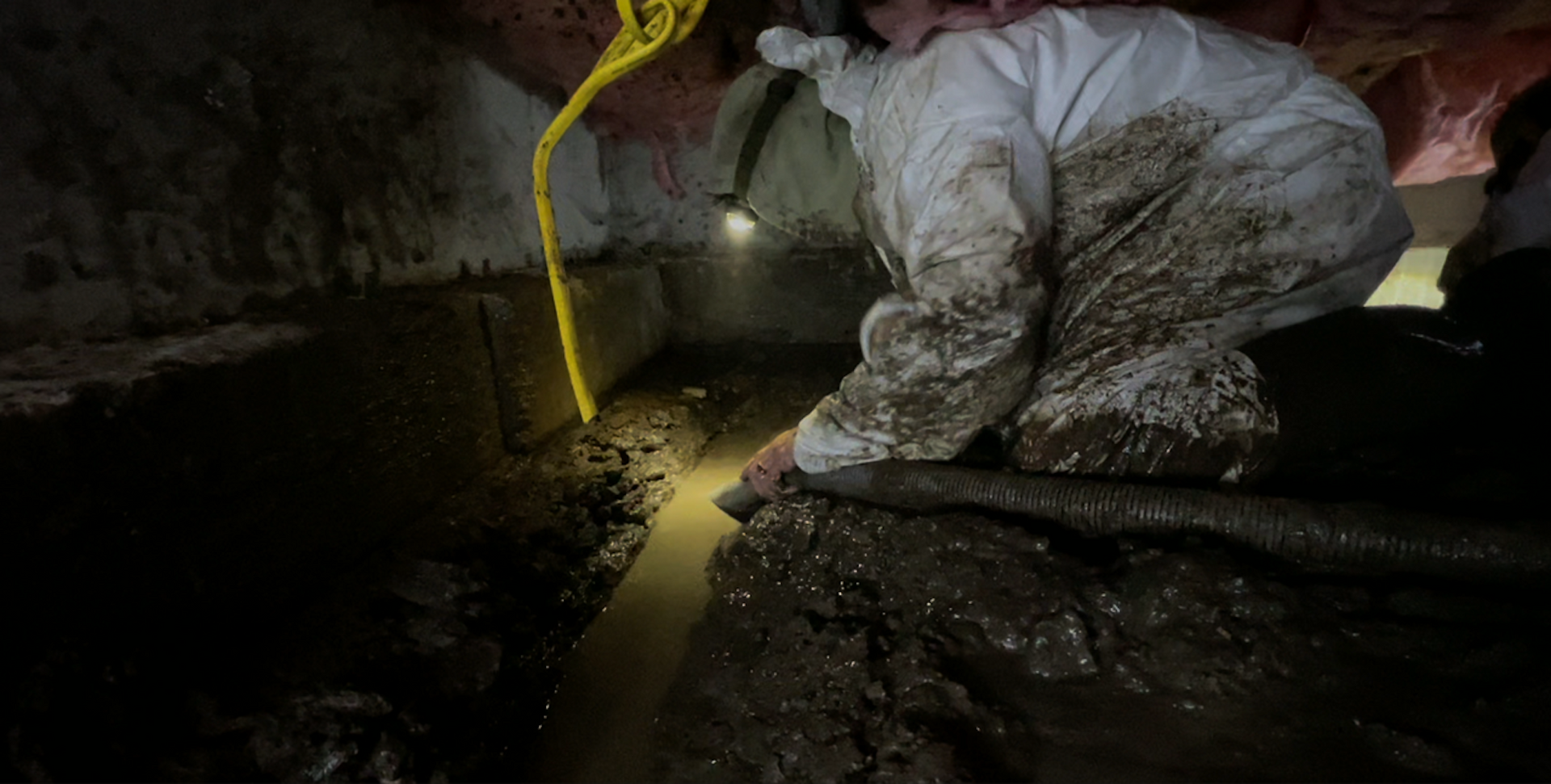 Worker in white protective suit kneels in a dark tunnel inspecting muddy water with a flashlight