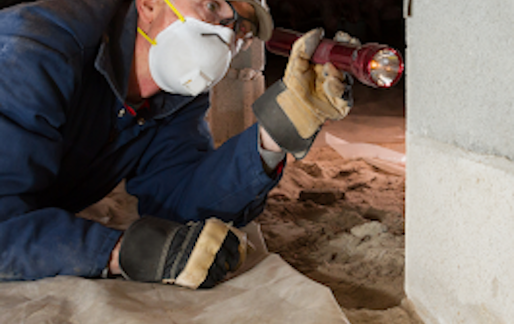 Worker in blue coveralls inspects insulation under a floor using a flashlight and mask