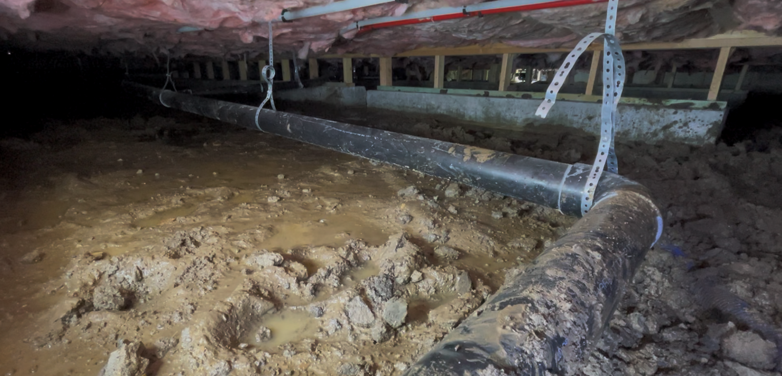 Flooded crawlspace under a house with muddy ground, exposed pipes, and beams overhead