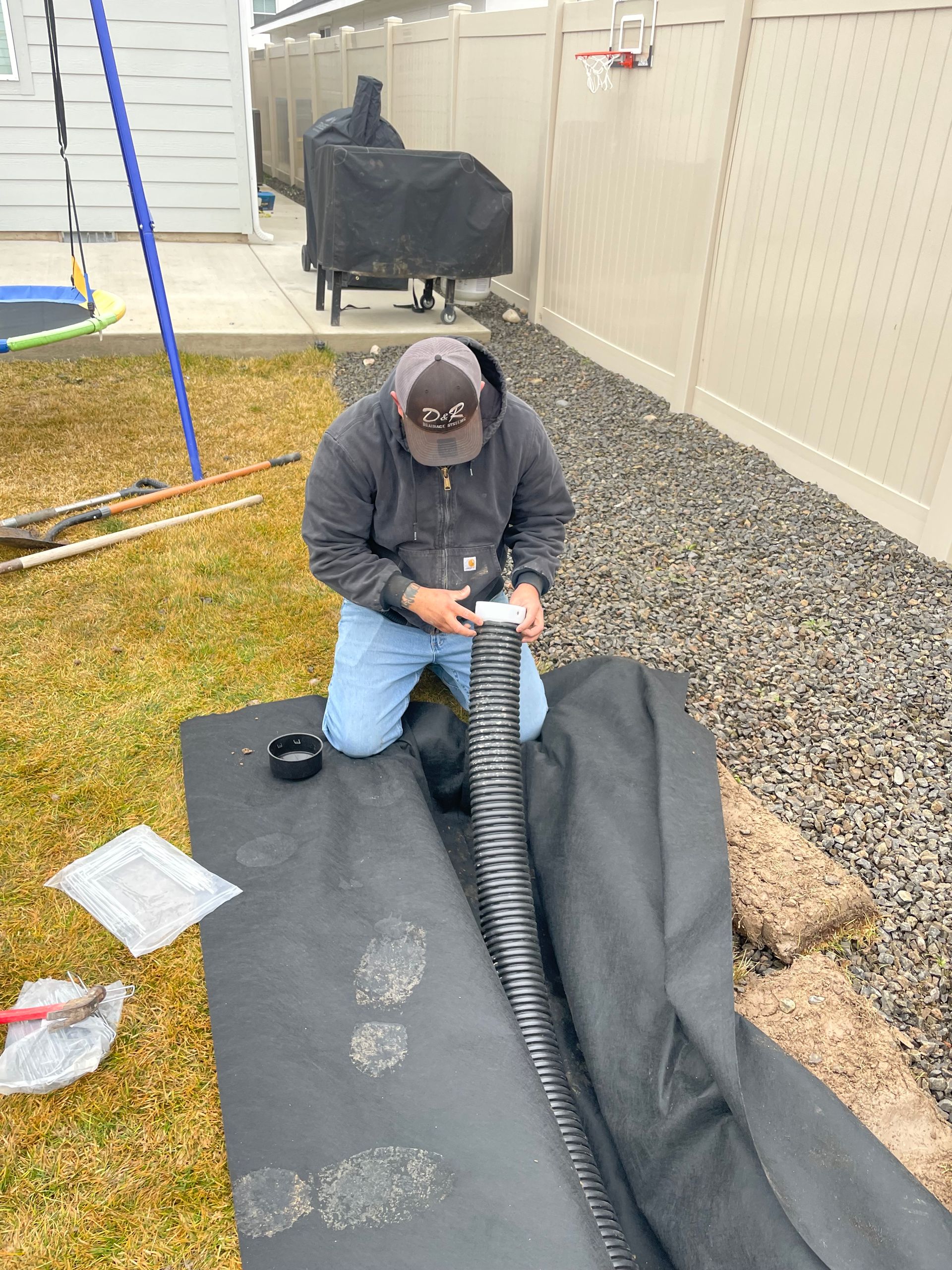 Person kneeling outdoors, securing black landscape fabric in a gravel yard beside a house.