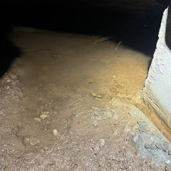 A pool of water sits on a dirt floor next to a concrete foundation wall in a dimly lit, enclosed crawl space.