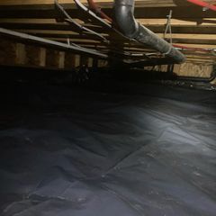 A dark crawl space with a black plastic vapor barrier covering the ground beneath wooden floor joists and plumbing pipes.