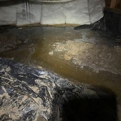 A flooded crawl space with standing water on a dirt floor, next to a black plastic vapor barrier and white insulation.