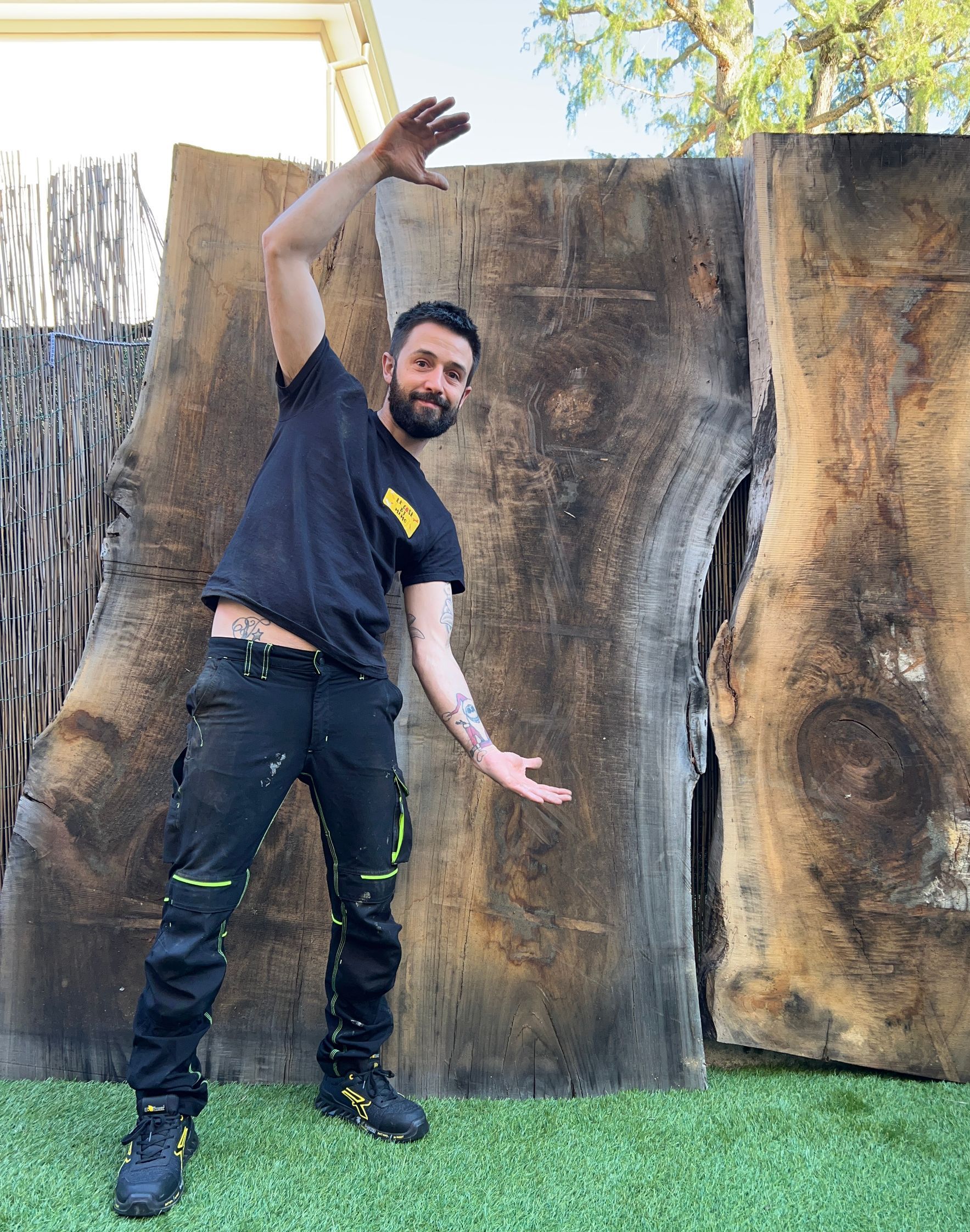 Un uomo con la barba indica due grandi lastre di legno grezzo sull'erba.