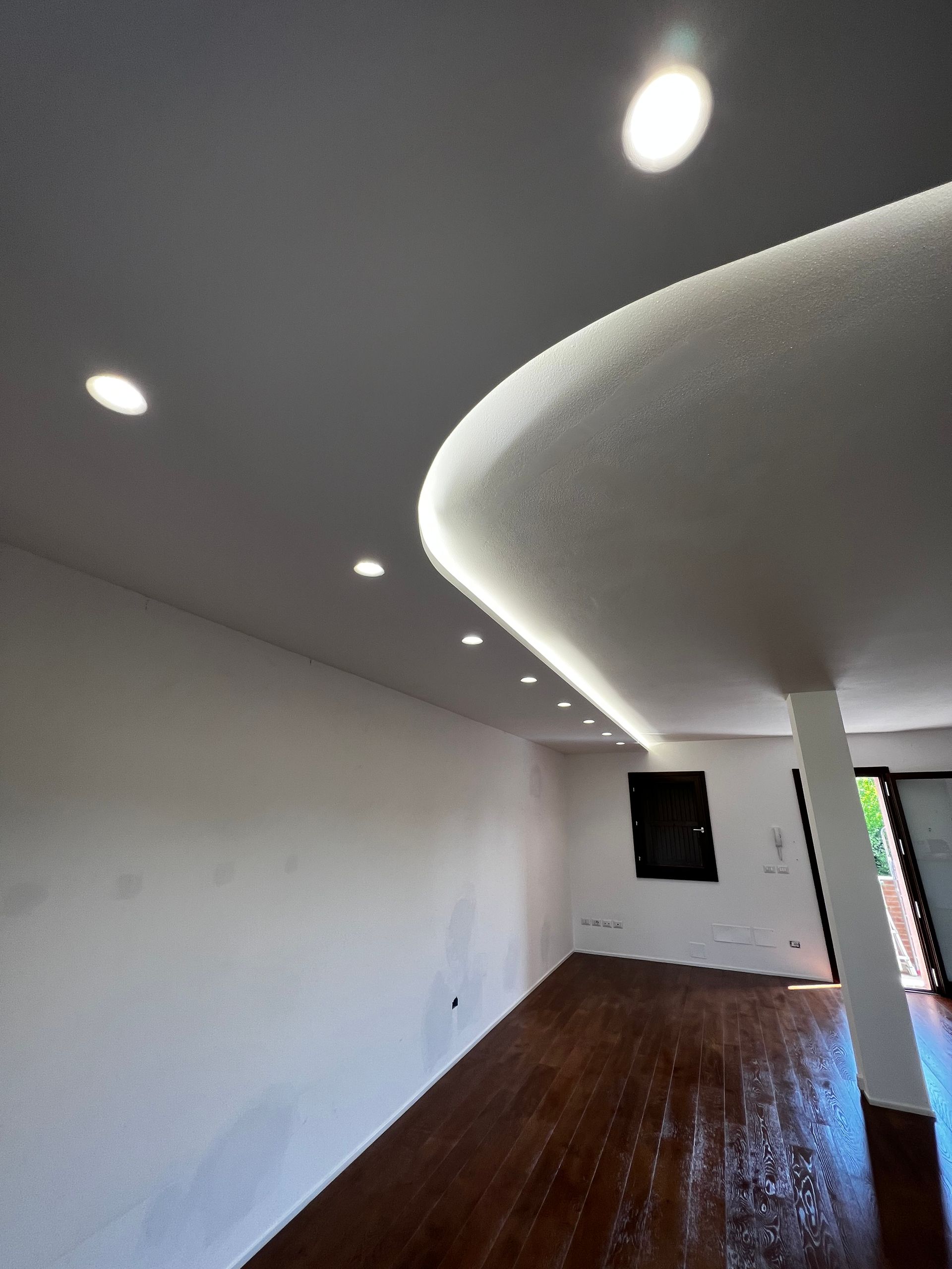 Vista interna di una stanza moderna con soffitto curvo e illuminazione da incasso. Pavimento in legno scuro, pareti bianche.