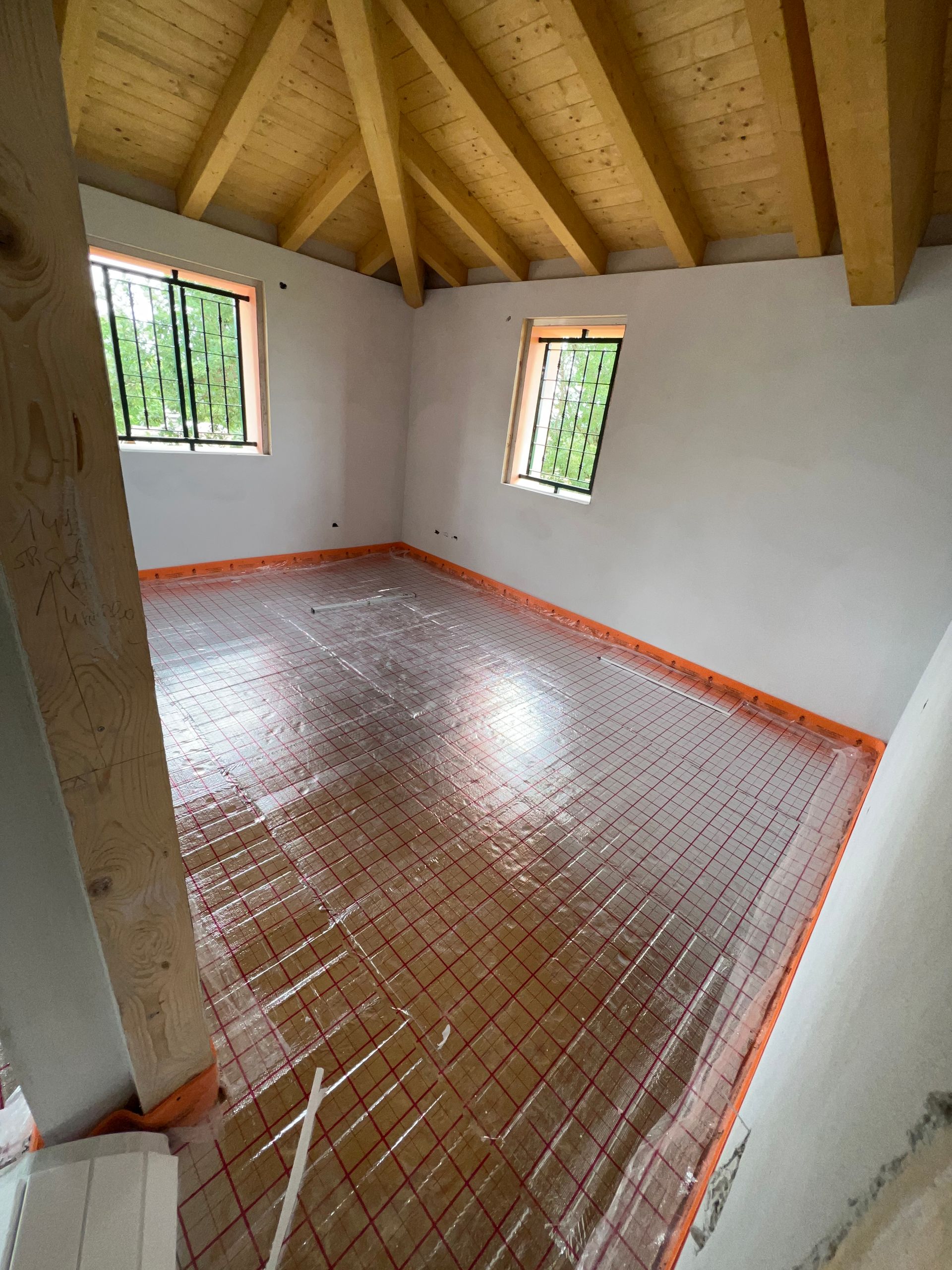 Stanza in costruzione con travi in ​​legno a vista, finestre e sistema di riscaldamento a pavimento radiante.