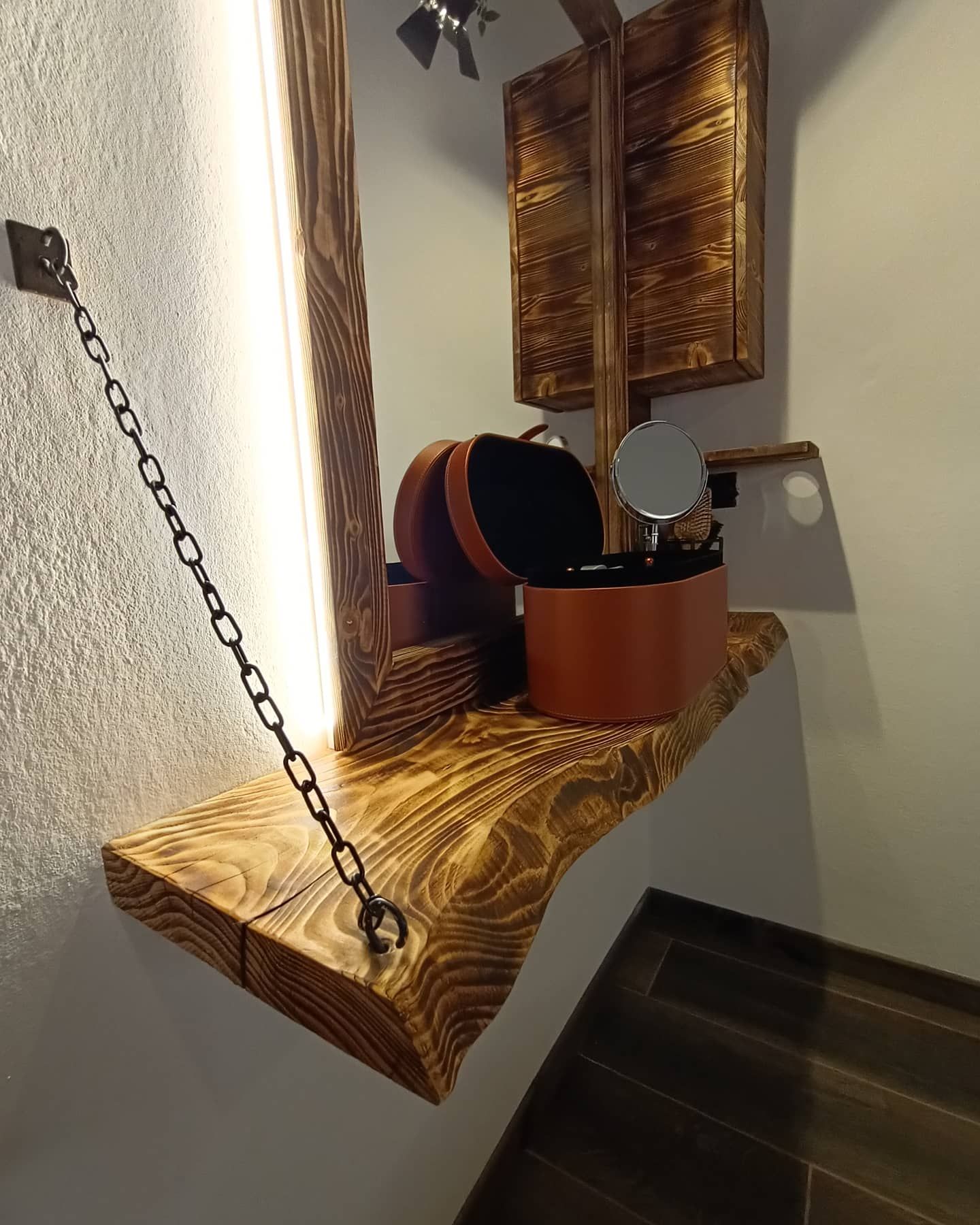 Mensola in legno con articoli per il trucco, illuminata da una striscia luminosa. Una catena sostiene la mensola, con un piccolo specchio e contenitori.