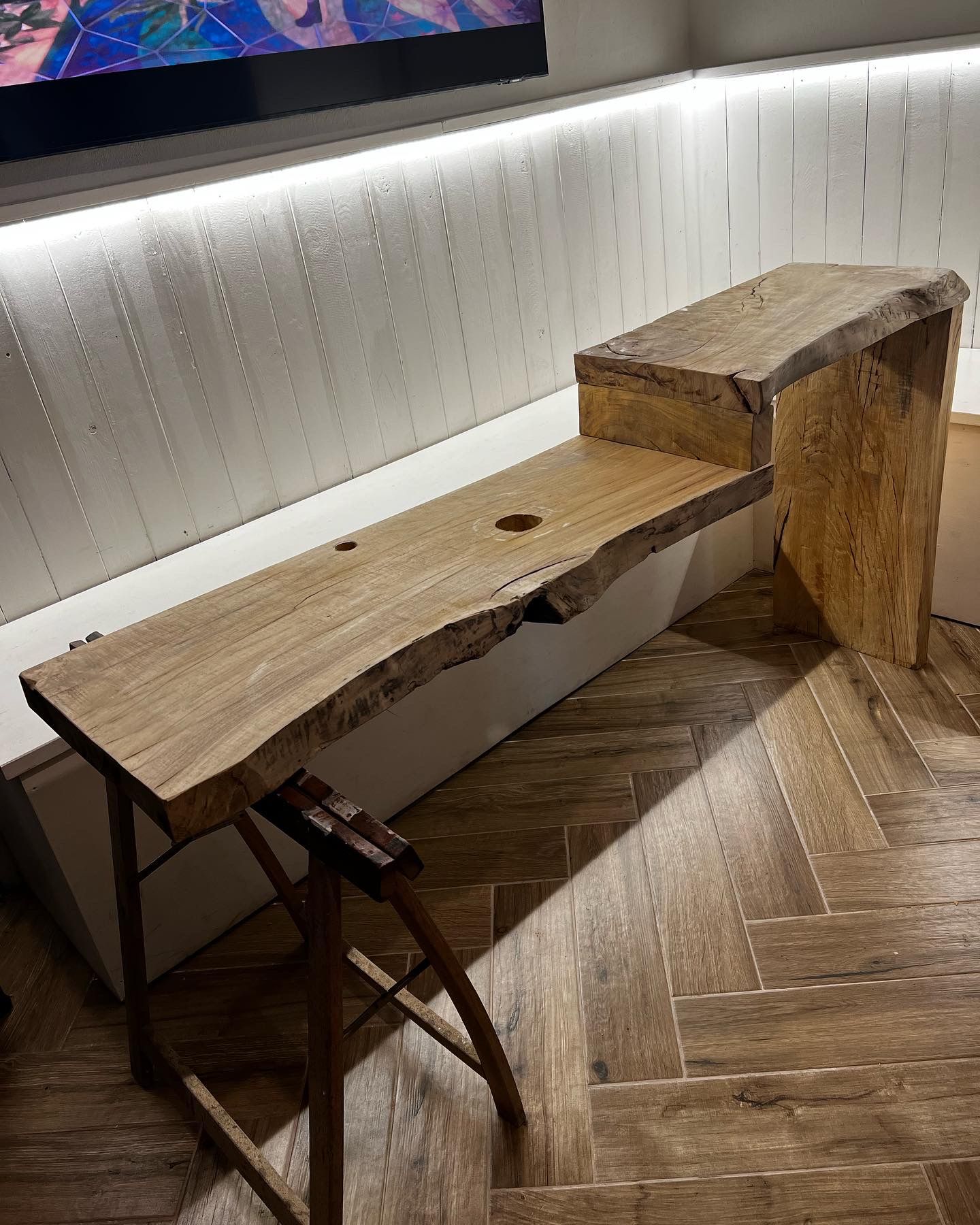 Scrivania in legno con bordo vivo e piccolo gradino; appoggiata su un basso ripiano bianco. Un'estremità poggia su un supporto pieghevole in legno.