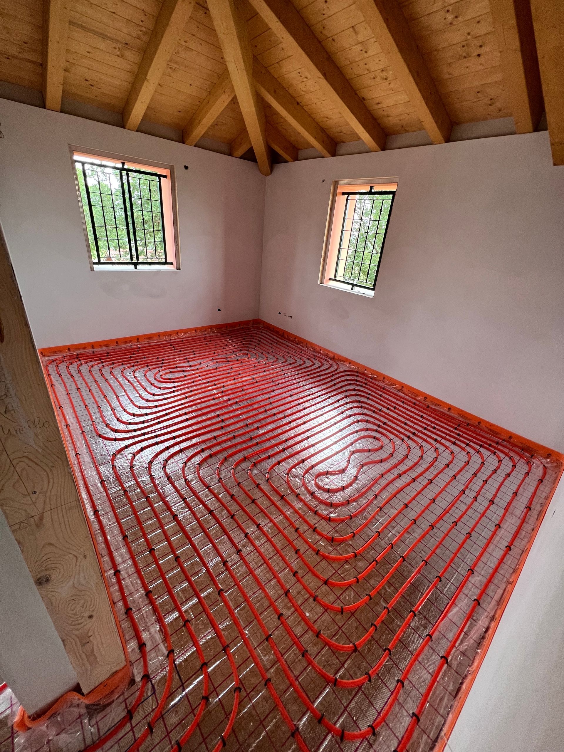 Stanza con sistema di riscaldamento a pavimento radiante rosso installato, finestre, soffitto in legno.