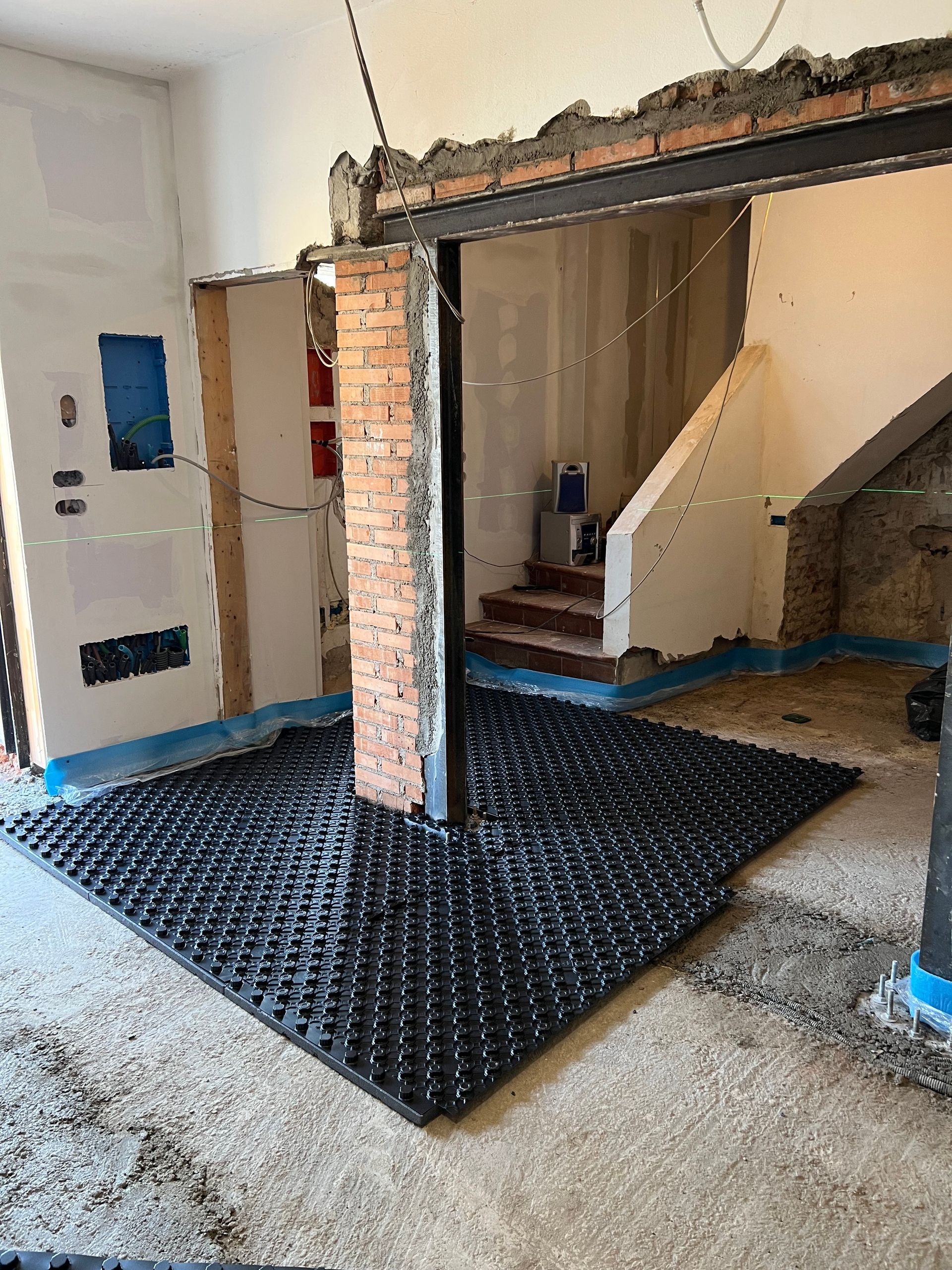 Cantiere interno con strato di drenaggio nero sul pavimento, pilastro in mattoni e scale sullo sfondo.