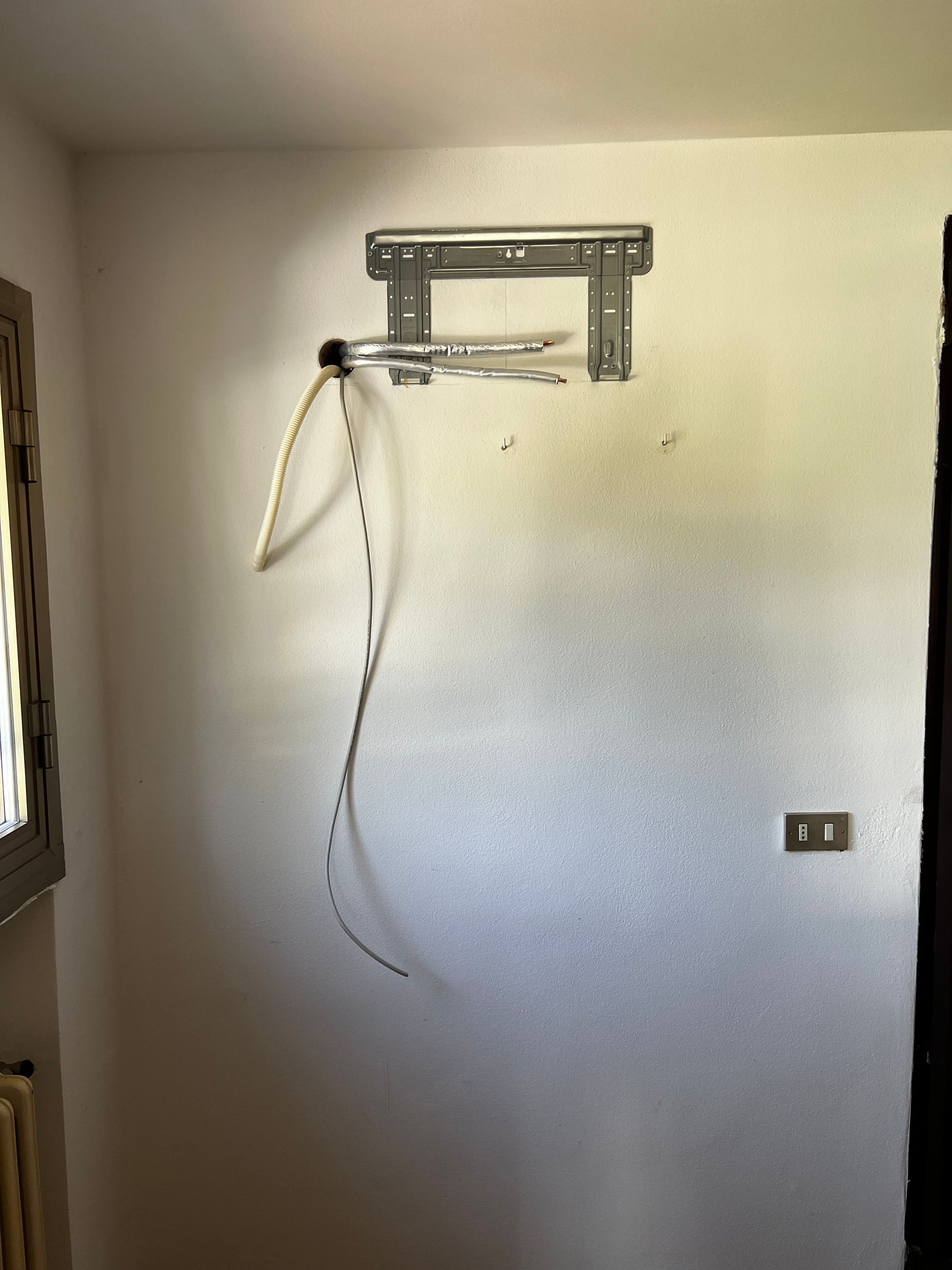 Supporto TV da parete con cavi pendenti; installato su una parete di colore chiaro con una presa elettrica e una porta.