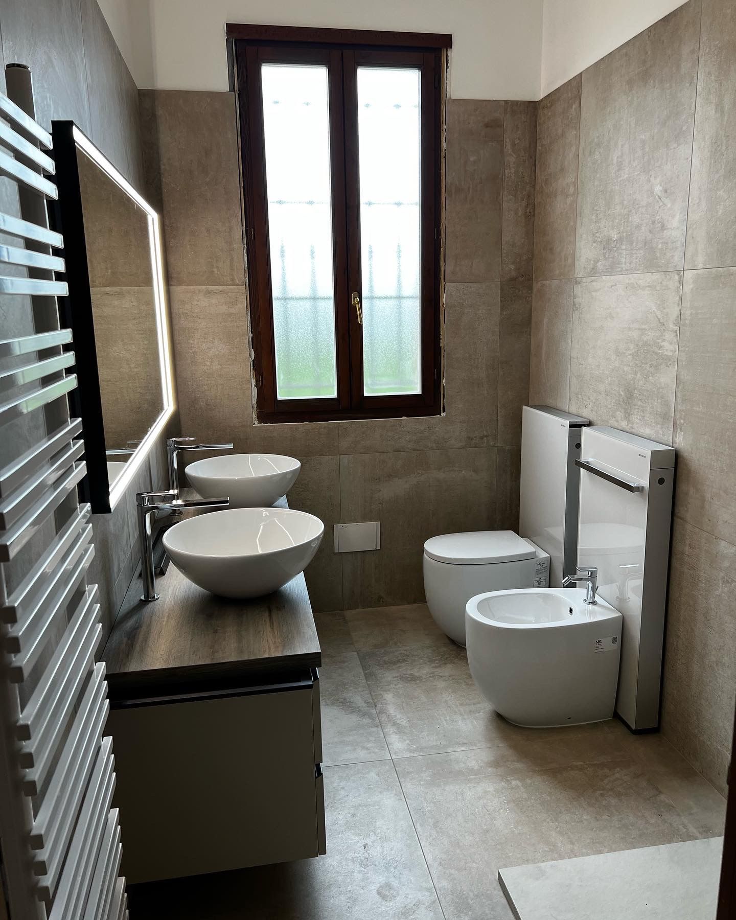 Bagno con due lavandini, WC e bidet. Finestra con cornice marrone. Pareti e pavimento piastrellati grigi.
