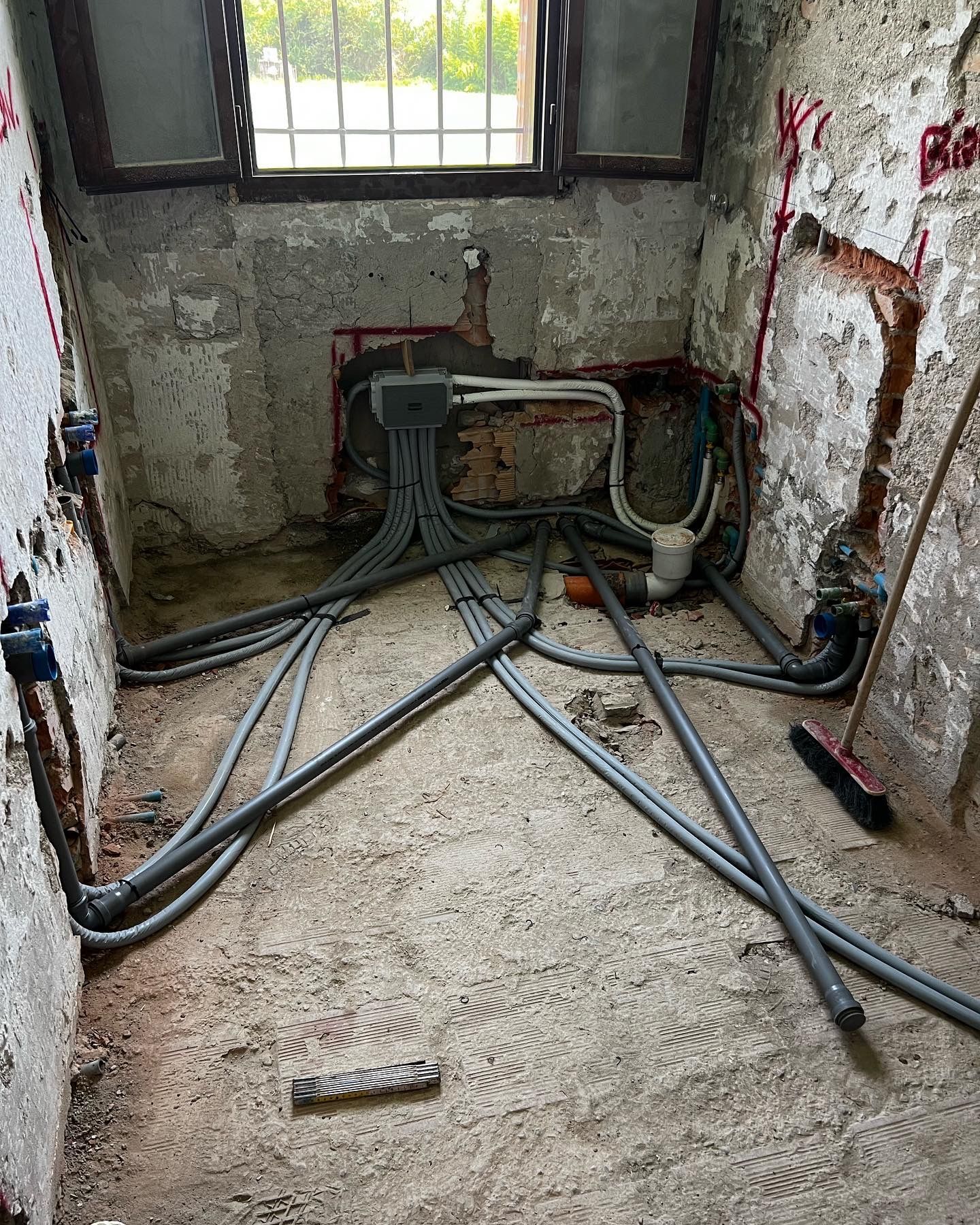 Bagno in costruzione: tubature e cavi elettrici a vista su pareti e pavimento in cemento. Finestra sullo sfondo.