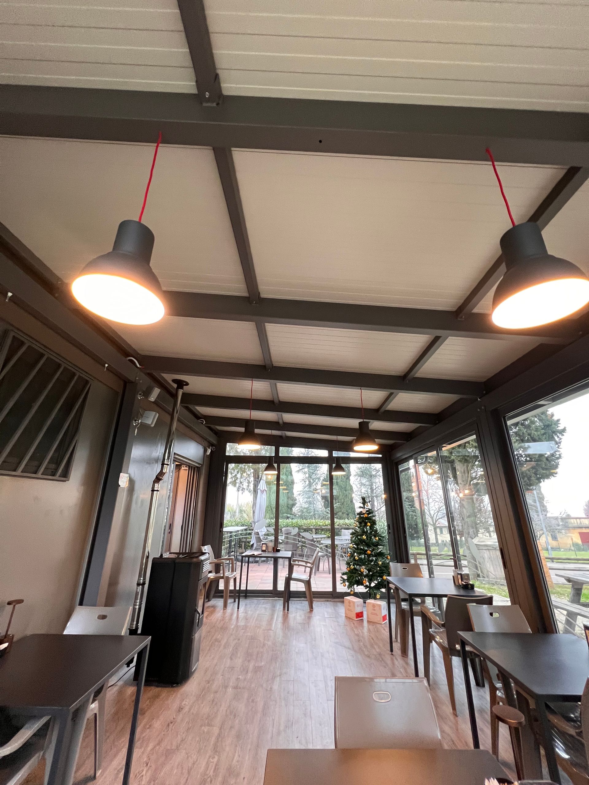 Bar all'aperto con tavoli e sedie, sotto una tettoia. Sono visibili lampade a sospensione e un piccolo albero di Natale.