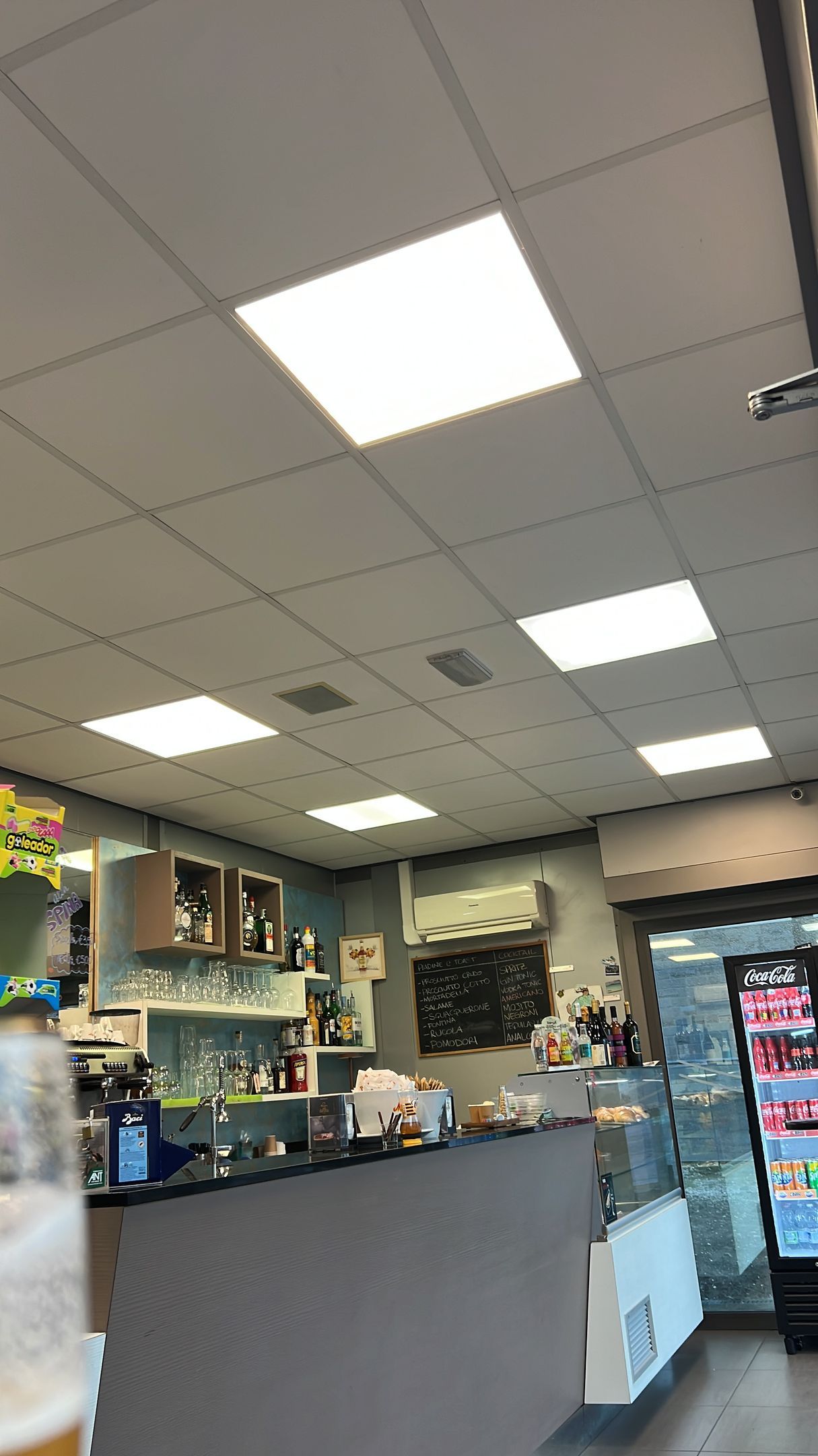 Interno di un bar con bancone, frigorifero espositivo e diverse luci sospese.